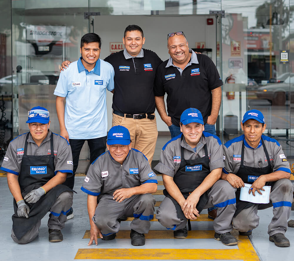 Grupo de hombres con camisas azules y chalecos negros, sonriendo y conversando en un ambiente amigable.